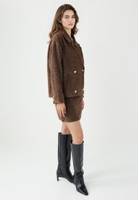 Bruine gestructureerde jas met dubbelzijdige gouden knopen, gecombineerd met een bijpassende rok en kniehoge zwarte leren laarzen.