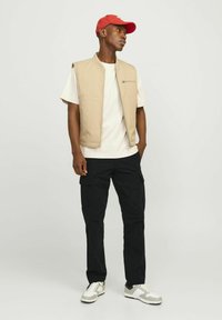 Vesta bej fără mâneci peste un tricou alb, combinată cu pantaloni cargo negri. Încălțările sunt adidași gri și albi. O șapcă roșie aduce un accent de culoare.