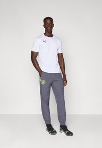 Hvid sports T-shirt med sort Puma-logo, parret med grå joggers med et grønt cirkulært emblem. Sorte sportssko fuldender outfittet.