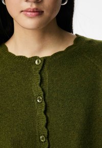 Zaļš adīts džemperis ar viļņotu izgriezumu, kurā ir piecas apaļas pogas nomierinošā tonī un mīksts, fakturēts audums.