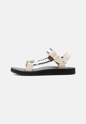 Beige og hvit sandal med åpen tå og justerbare stropper, med en svart teksturert såle, sidevisning på en ensfarget lys bakgrunn.