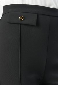 Close-up of black high-waist pants with a fabric flap and round black-and-gold button detail at the waistband.