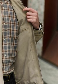 Chaqueta beige con una textura suave, desabrochada para revelar una camisa de cuadros en azul, marrón y crema. Cuenta con un bolsillo lateral y cierre de botones.