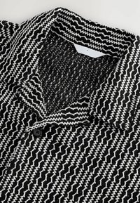 Collar de camisa de patrón en blanco y negro que presenta un tejido texturizado, detalle de botones y líneas en zigzag repetidas en colores contrastantes.