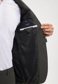 Mężczyzna w białą w paski koszuli pokazuje czarną wzorzystą podszewkę oliwkowego, dopasowanego garnituru JOOP!