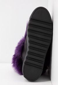 Černé tenisky s texturovanou gumovou podrážkou a fialovým umělým kožíškem, nakloněné proti bílému pozadí, ukazující svou unikátní konstrukci.