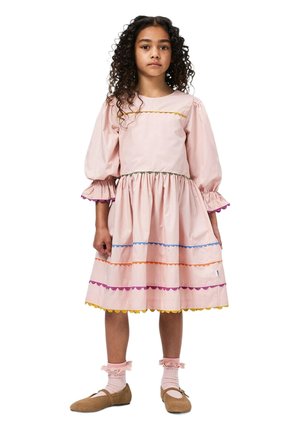 Vestido de algodón rosa claro con mangas abullonadas, cintura fruncida y ribete ondulado colorido en amarillo, naranja, azul y morado. Zapatos planos marrones.