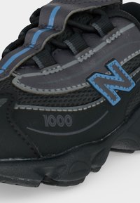 Μαύρο και γκρι αθλητικό παπούτσι New Balance με μπλε ρίγα, λογότυπο "N", κείμενο "1000" και μαύρα κορδόνια σε υφασμένο πάτο.