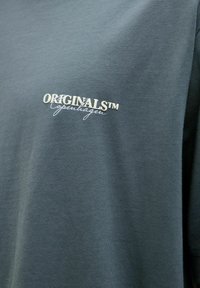 Σκούρο γκρι-πράσινο ύφασμα με το κείμενο «ORIGINALS™ Copenhagen» τυπωμένο σε λευκό στην περιοχή του στήθους ενός ενδύματος.