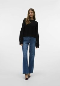 Zwarte gebreide trui met lange mouwen, relaxed fit, gecombineerd met uitlopende blauwe jeans. Zilveren accenten en puntige patenten hakken maken de look compleet.