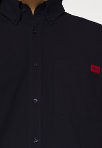 Camisa negra de botones con un collar estructurado, bolsillo frontal y una pequeña etiqueta de logo roja. La tela parece suave y duradera.