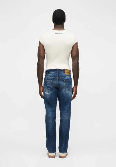 Homme debout de dos, portant un t-shirt blanc ajusté et un jean bleu déchiré avec un patch en cuir marron clair à la taille.