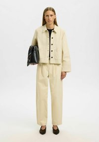 Bawełniana kurtka w kolorze kremowym z czarnymi guzikami i dwoma kieszeniami, zestawiona z pasującymi szerokimi spodniami i czarną torebką z kolorowym wzorem.