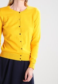 Gul stickad kofta med rund hals, guldfärgade knappar framtill och ribbade muddar. Mjuk textur, figurnära passform, kombinerad med en mörk kjol.