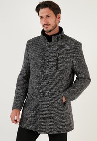 Abrigo de patrón gris y negro con un alto cuello, cinco botones y un bolsillo con cremallera. Presenta un material texturizado similar a la lana.