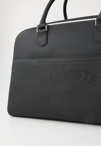 Borsa sintetica nera con superficie testurizzata, accenti bianchi, manici superiori, logo in rilievo e grafica di coccodrillo. Design strutturato con dettagli minimi.