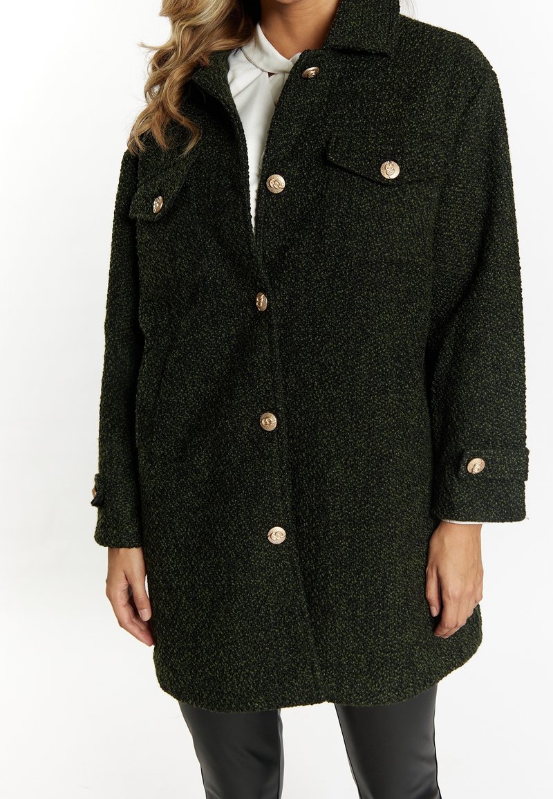 Cappotto bouclé verde con ampie tasche frontali, bottoni dorati e polsini. La texture è calda e strutturata, progettata con colletto.