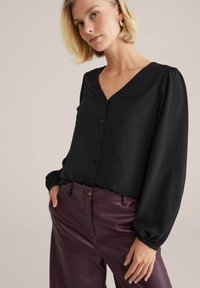 Blusa nera con scollo a V, chiusura a bottoni e maniche a sbuffo. Abbinata a pantaloni in pelle bordeaux, che mostrano una texture liscia.