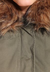 Olivgrön jacka med en brun faux fur-krage. Tyget är strukturerat och tjockt, med synliga stygn längs framsidan.