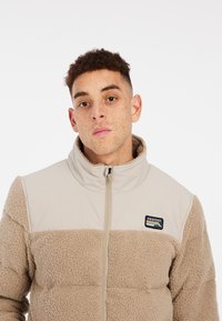 Veste en polaire beige avec fermeture éclair, col montant et partie supérieure lisse. Présente une étiquette noire avec "PROTEST" et un motif de montagne.