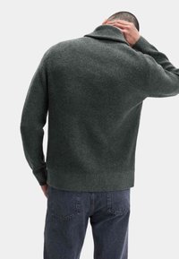 Mørkegrøn strikket sweater med høj krave, ribbet tekstur og lange ærmer, båret med mørke jeans, set fra bagsiden.