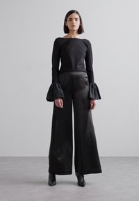 Černý top s dlouhým rukávem a zvonovými rukávy, spárovaný s kalhotami s širokými nohavicemi. Hladká tkanina s matným povrchem, minimalistický design. Černé kotníkové boty.