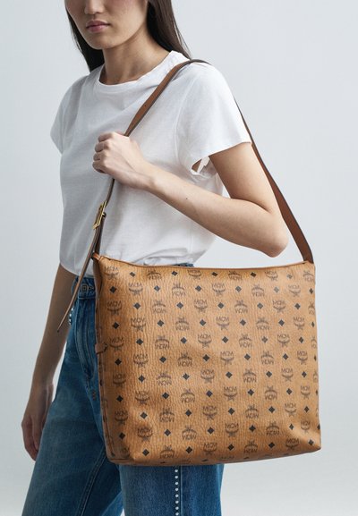 Brązowa skórzana torba na ramię z tłoczonymi wzorami logo, gładką fakturą i regulowanym brązowym paskiem, trzymana przez osobę w dżinsach.