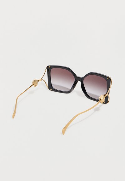 Lunettes de soleil carrées noires avec des verres dégradés et des branches fines dorées ornées de petits détails de têtes de lion près des charnières.