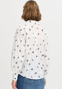 Weiße Bluse mit navyfarbener Blattmuster, langen Puffärmeln, einem Knopfkragen und einem leicht abgerundeten Saum. Leichte, leicht durchsichtige Stoff.