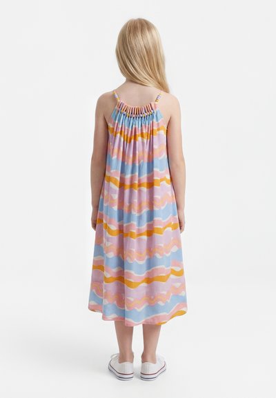 Νεαρή κοπέλα με μακριά ξανθά μαλλιά, φορώντας ένα αμάνικο, πολύχρωμο φόρεμα με κυματιστές ρίγες και λευκά αθλητικά, στέκεται με την πλάτη προς τα εμπρός.