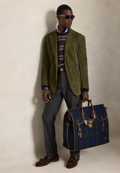 Uomo con occhiali da sole, blazer verde, maglione fantasia, pantaloni grigi e mocassini marroni che tiene una grande borsa a quadri blu e verde con rifiniture in pelle.