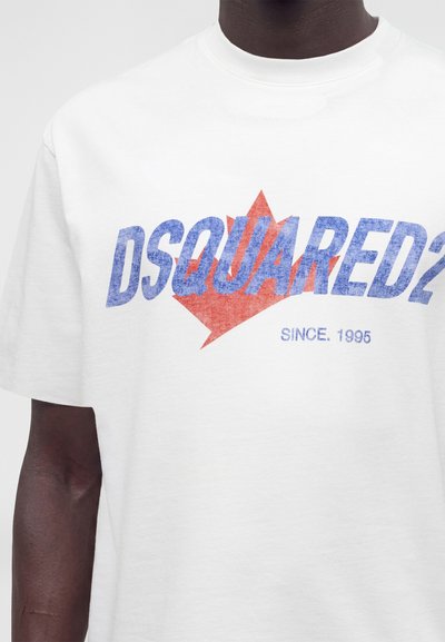 Λευκό μπλουζάκι με μπλε κείμενο "DSQUARED2" πάνω από κόκκινο γραφικό σχήμα φύλλου σφενδάμου και μικρότερο μπλε κείμενο που γράφει "Since 1995" στο στήθος.