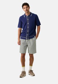 Marineblauwe korte mouwen overhemd met witte bies, gecombineerd met lichtgestreepte shorts. Het model draagt beige sneakers en witte sokken met gele strepen.