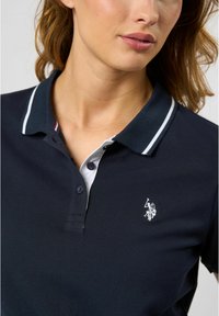 Marineblå polo t-shirt lavet af bomuld, med en hvid krave med rød og blå striber, to knapper og et lille hvidt logo på brystet.
