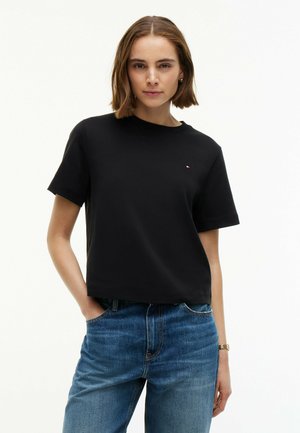 Jonge vrouw met kort bruin haar die een effen zwart T-shirt en blauwe spijkerbroek draagt, staand met één hand in de zak tegen een witte achtergrond.