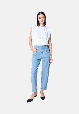 Jeune femme debout portant un haut blanc sans manches, un jean taille haute bleu clair et des talons pointus noirs, devant un fond uni.