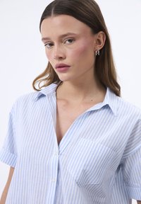 Camicia a righe azzurro chiaro e bianco con colletto, maniche corte e taschino sul petto; realizzata in un tessuto morbido e leggero. Chiusura frontale con bottoni.