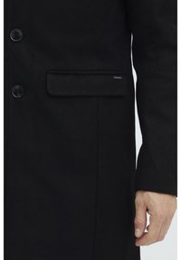 Schwarzer Wollmantel mit drei Knöpfen, einer einzelnen Fronttasche und strukturierter Stoff. Knopfdetails sind sichtbar; minimalistisches Design.