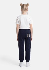 Copil cu păr blond, aranjat în două cocuri și codițe laterale, purtând un tricou alb, pantaloni sport bleumarin cu patch „II” și pantofi sport albi, cu fața spre spate.