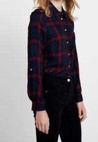 Marineblauw geruit overhemd met rode lijnen, met knopen, lange mouwen. Gouden knoopaccenten op de manchetten, gemaakt van een zachte stof.