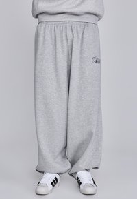 Graue, lockere Sweatpants aus weichem Stoff mit elastischem Bund. Mit gesticktem Detail am Oberschenkel und kombiniert mit weißen Turnschuhen.