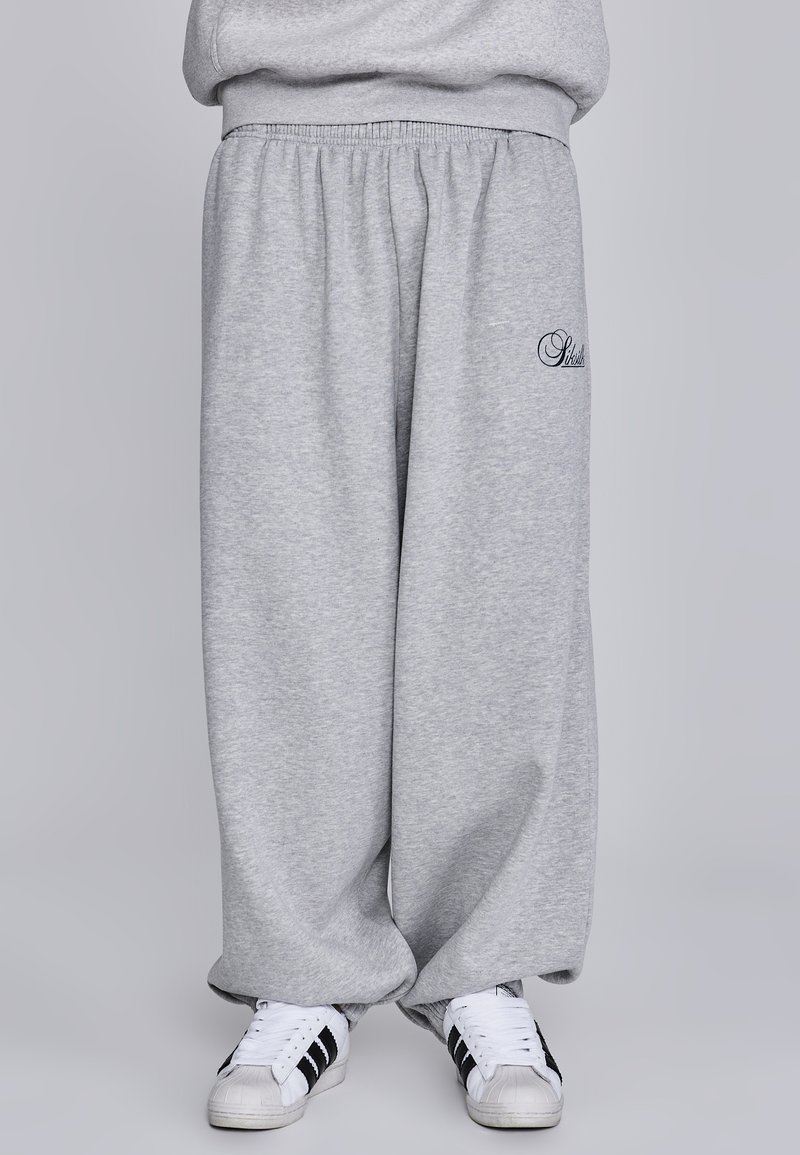 Graue, lockere Sweatpants aus weichem Stoff mit elastischem Bund. Mit gesticktem Detail am Oberschenkel und kombiniert mit weißen Turnschuhen.
