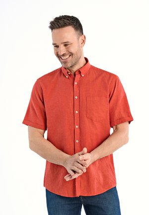 Man glimlachend, draagt een rood overhemd met korte mouwen en blauwe spijkerbroek, met handen gevouwen voor zich, staand tegen een witte achtergrond.