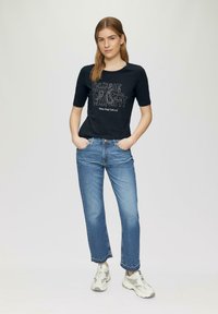 T-shirt en coton noir avec un col rond et des manches courtes, arborant un texte graphique blanc. Porté avec un jean bleu clair et des baskets grises.