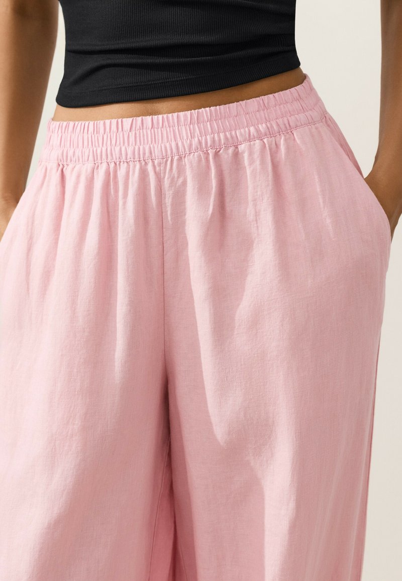 Femme portant un pantalon rose fluide à taille haute avec une ceinture élastique et des poches avant, assorti à un top court noir.