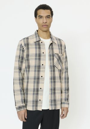 Mand iført beige og marineblå ternet skjorte med knapper over en hvid t-shirt og sorte bukser, stående mod en ensfarvet hvid baggrund.