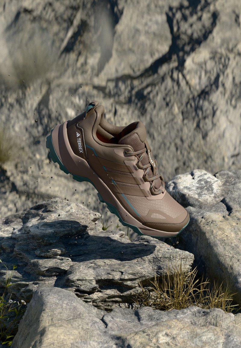Brauner Sportschuh mit strukturiertem Obermaterial, türkisfarbenen Akzenten und gemusterter Sohle, in der Luft über steinigem Terrain positioniert.