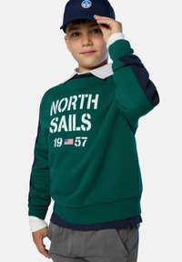 Sudadera verde con el texto "NORTH SAILS 1957", detalles en blanco y mangas en azul marino. Lleva una camiseta azul marino con un cuello blanco por debajo.