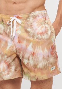 Badehosen mit einem Batikmuster in pastellfarbenen Tönen, ausgestattet mit einem elastischen Bund, einem weißen Zugband und einem kleinen Markenetikett an der Seite.