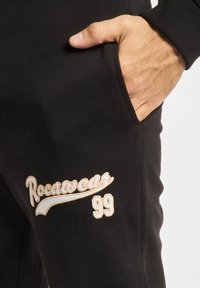 Melni sporta bikses ar gludu tekstūru, ar baltu un zeltītu "Rocawear" izšuvumu un skaitli "99". Redzama sānu kabatas detaļa.